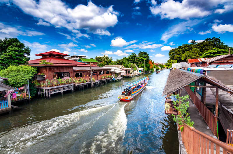 Barca tradicional navegando por los canales de Bangkok entre casas de madera y vegetación en la zona de Thonburi
